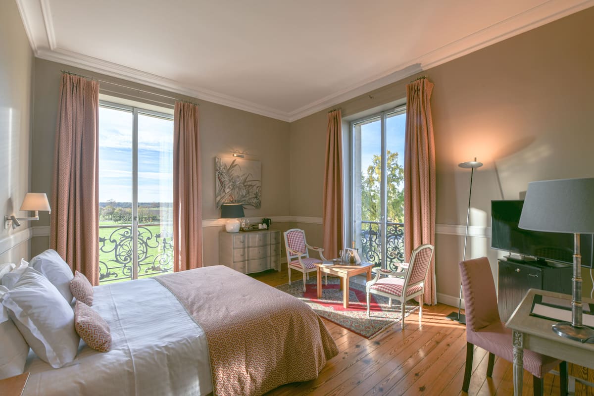 A hotel room with a large bed, a desk with a TV, a chair, and a balcony with a view.