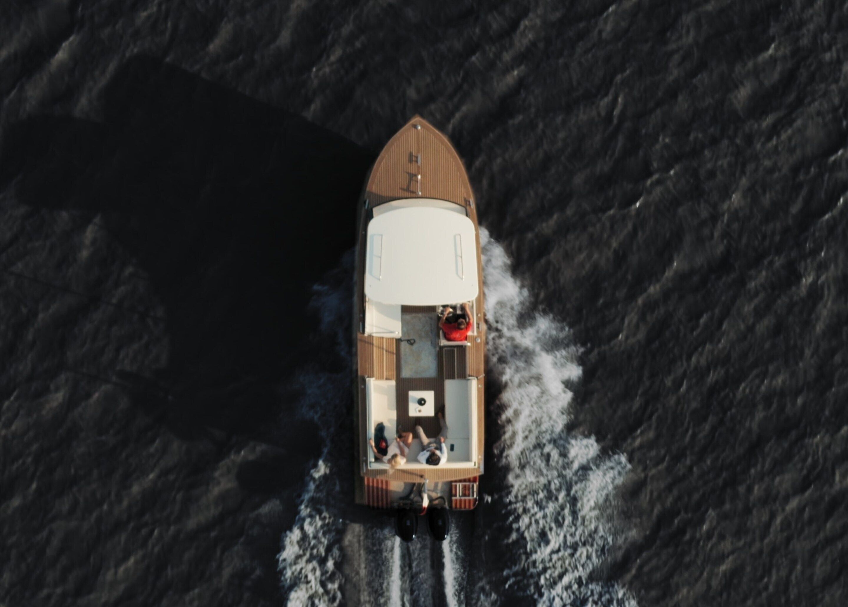 Vista aérea de un barco con cubierta de madera, un toldo blanco y dos personas sentadas al frente.