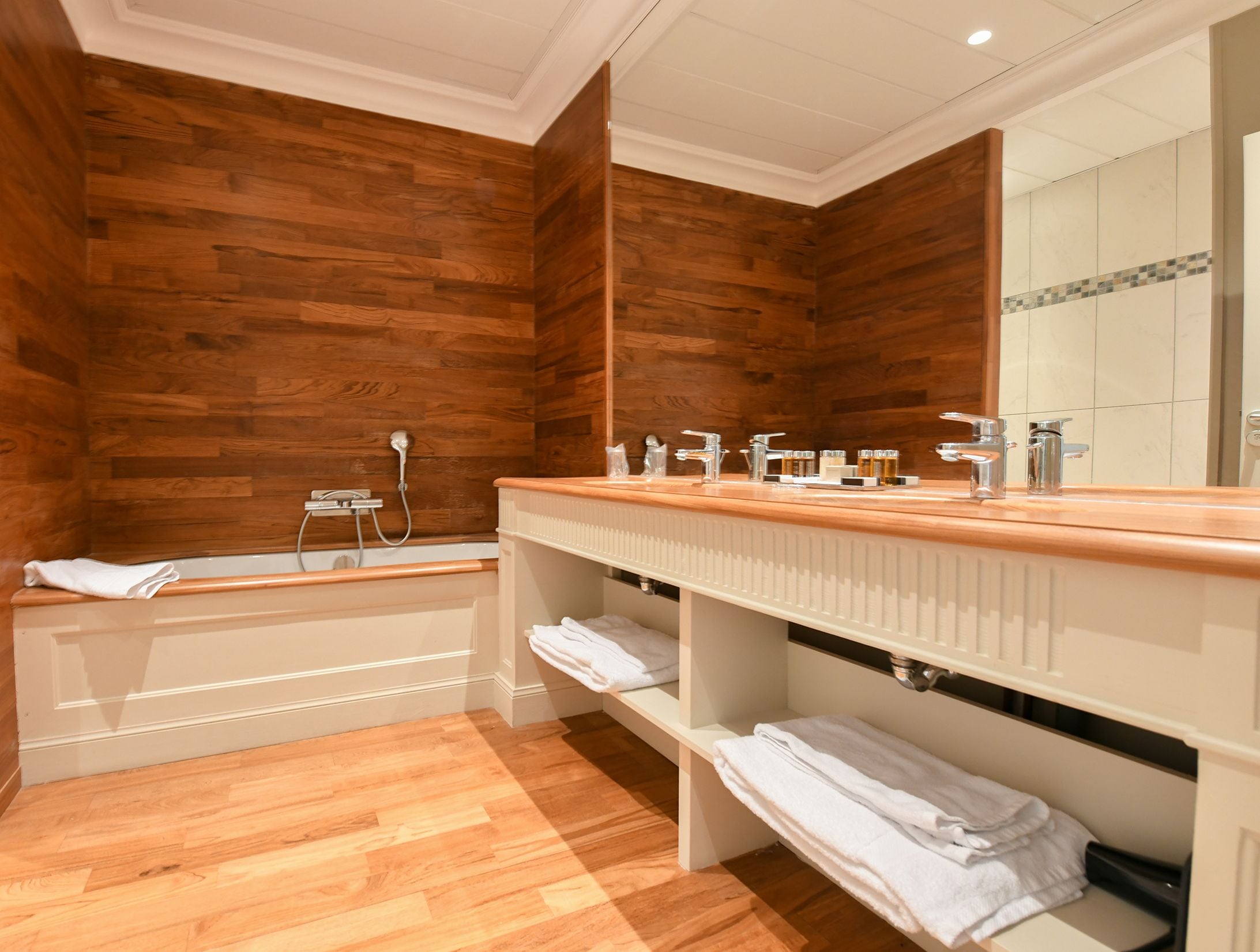 A bathroom with wooden panel walls, a wooden vanity with a mirror, a bathtub, and shelves with towels.