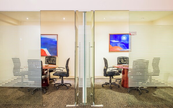 A conference room with glass partitions, two desks, black office chairs, and abstract paintings on the walls.