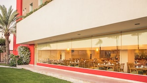 A modern building with a red exterior and a large glass facade. Inside, there are multiple dining tables and chairs arranged in a spacious, open layout. - Fiesta Inn Tijuana Otay (Tijuana)