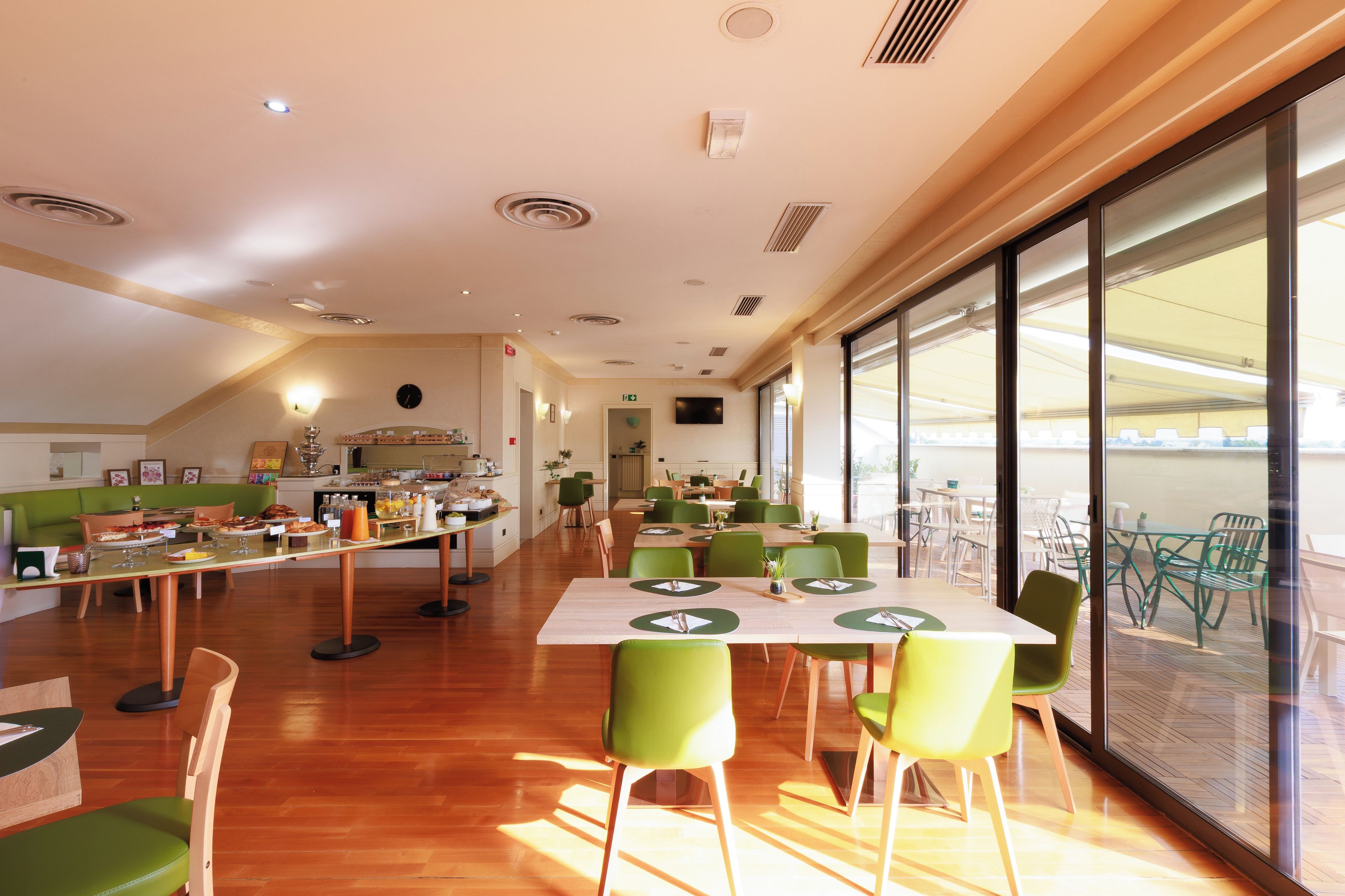 Un grand espace à manger avec plusieurs tables et chaises, une cuisine à l’arrière-plan et de grandes fenêtres qui laissent entrer la lumière naturelle.