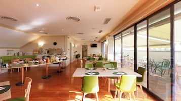 A spacious dining area with multiple tables and chairs, a kitchen in the background, and large windows providing natural light.