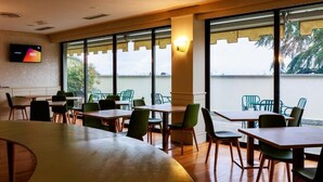 A modern dining area with wooden tables and green chairs, large windows with a view of palm trees, and a flat-screen TV mounted on the wall. - Hotel Excelsior Magenta (Magenta)
