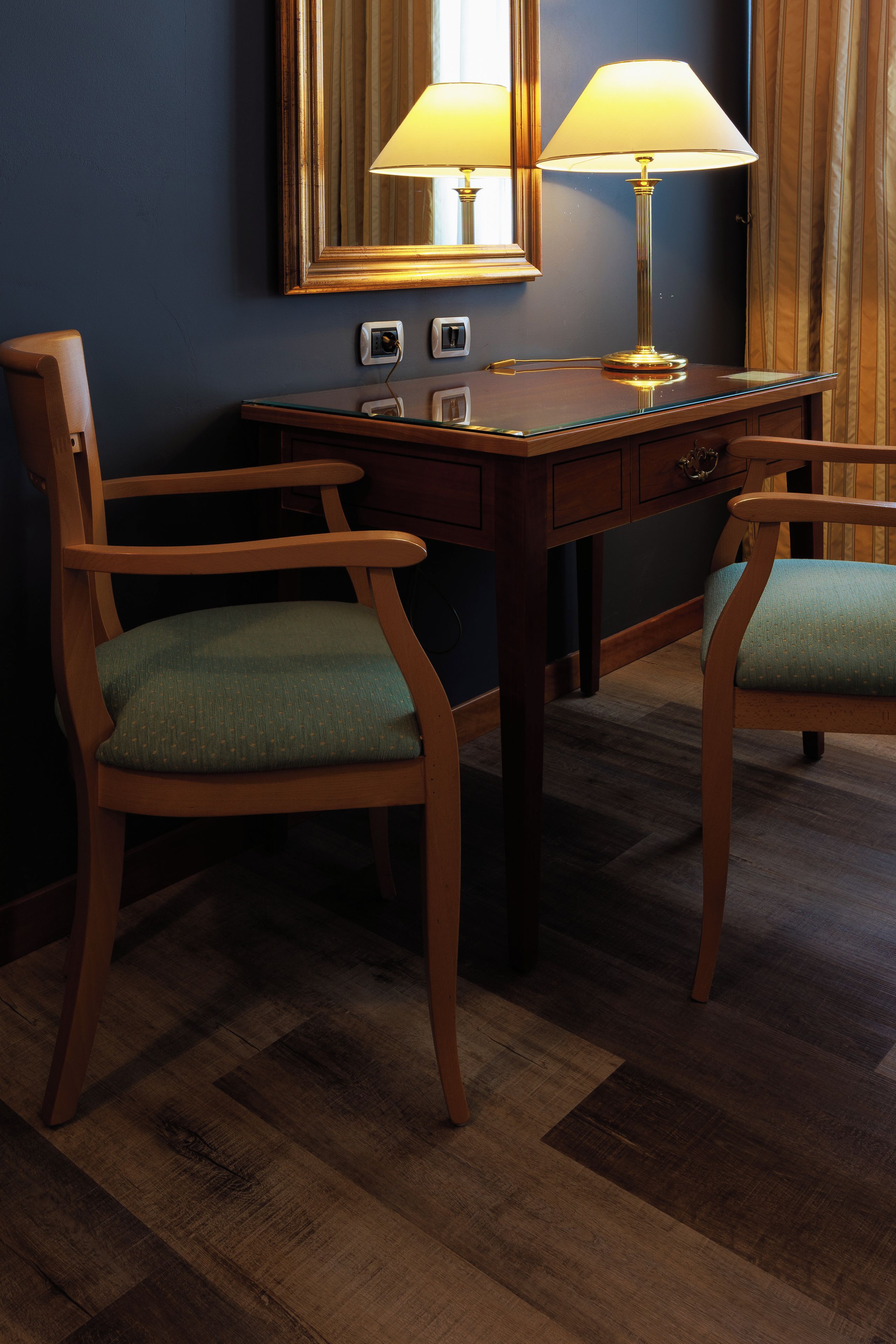 A hotel room with a wooden desk, two chairs, a lamp, and a mirror.