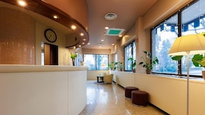A hotel lobby with a curved reception desk, a clock, potted plants, and large windows with natural light. - Hotel Excelsior Magenta (Magenta)