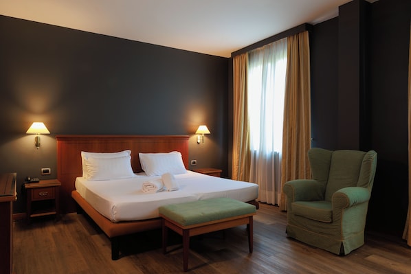 Una habitación de hotel con una cama grande, dos lámparas de noche, un sillón verde y una mesita de noche de madera.