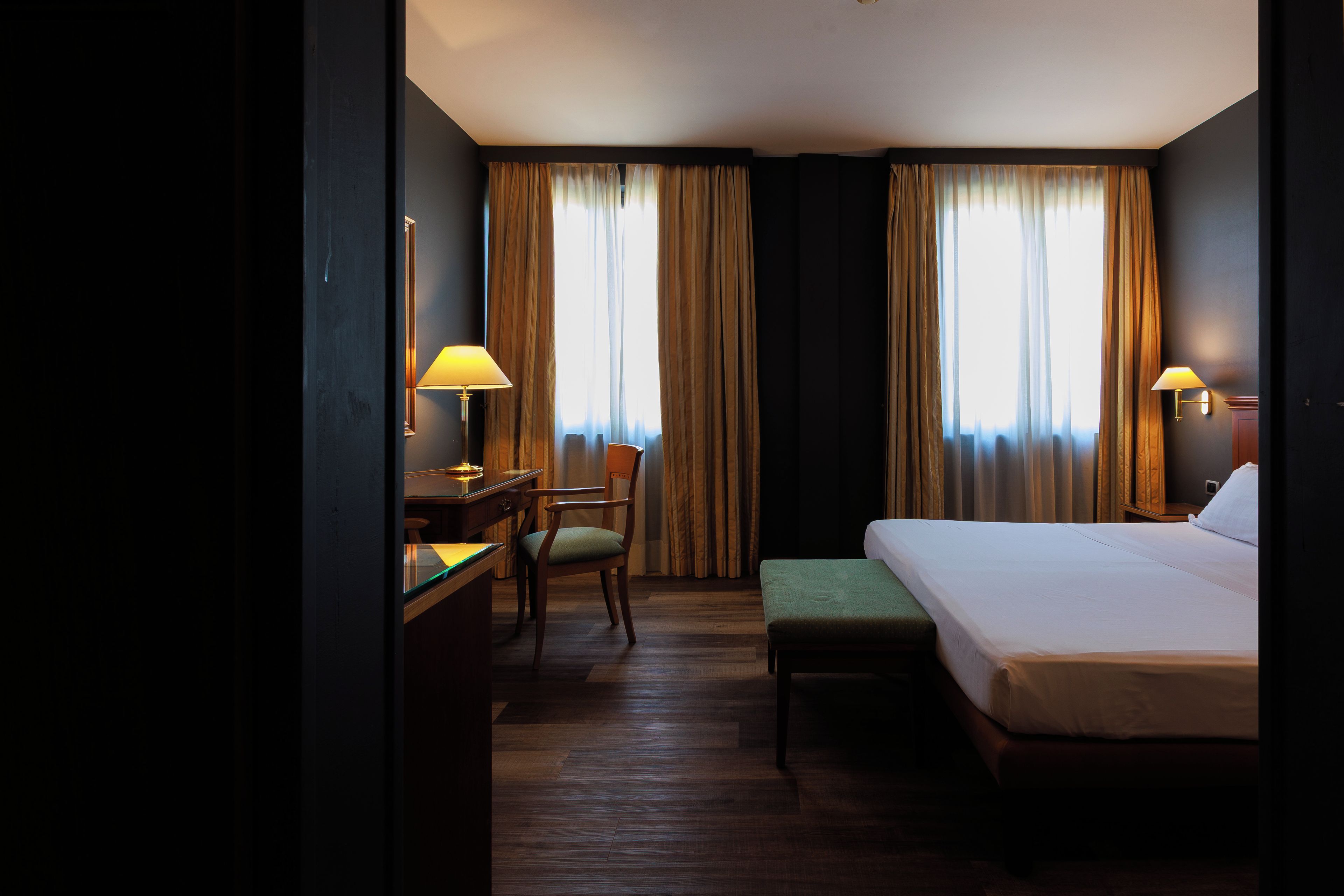 Habitació d'hotel amb un llit, una taula de treball, una cadira i dues finestres amb cortines.