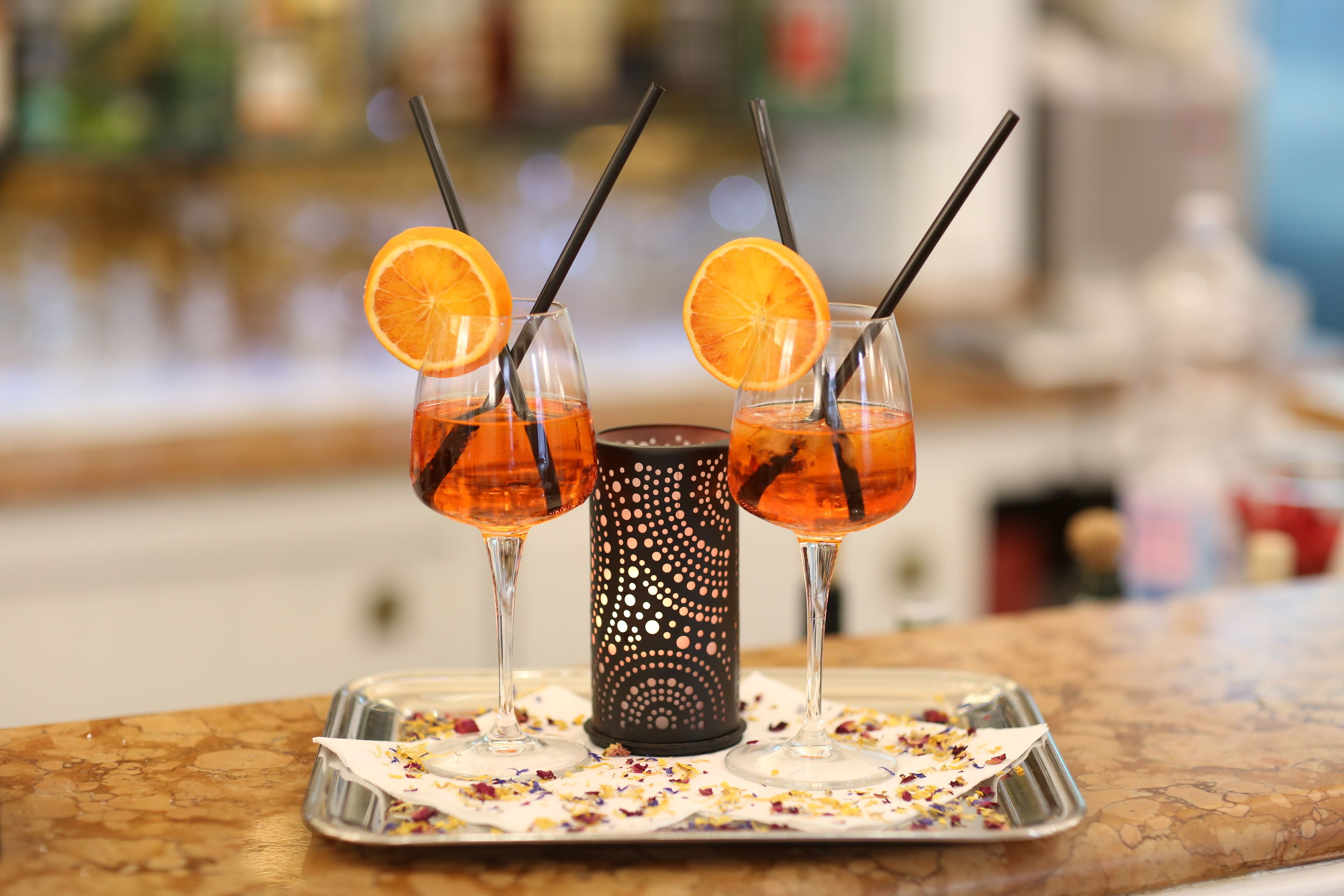 Three glasses of orange-colored beverage with straws, a decorative candle, and a tray with confetti on a marble surface.