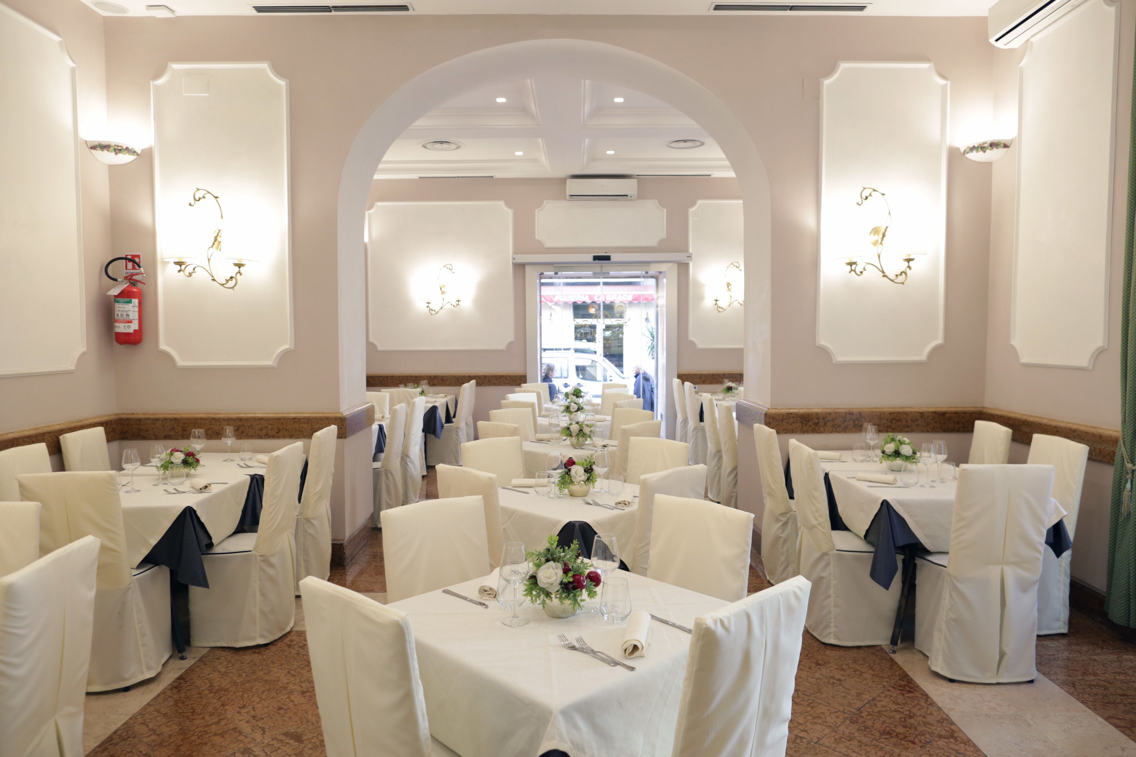 A banquet hall with multiple round tables, each set with white tablecloths and chairs, and a central aisle leading to a door.