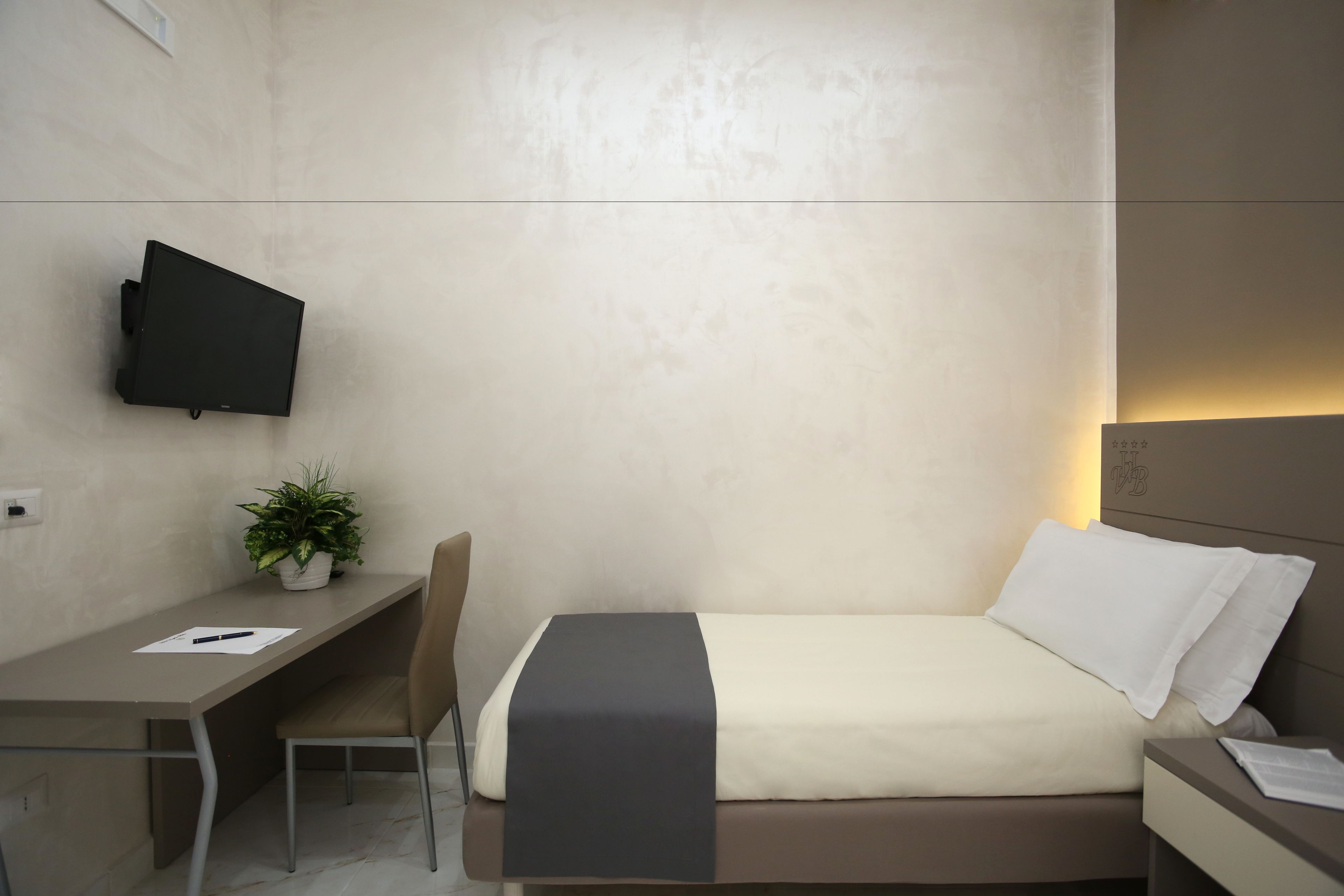 A modern hotel room with a bed, a desk, a chair, a TV mounted on the wall, and a small potted plant.