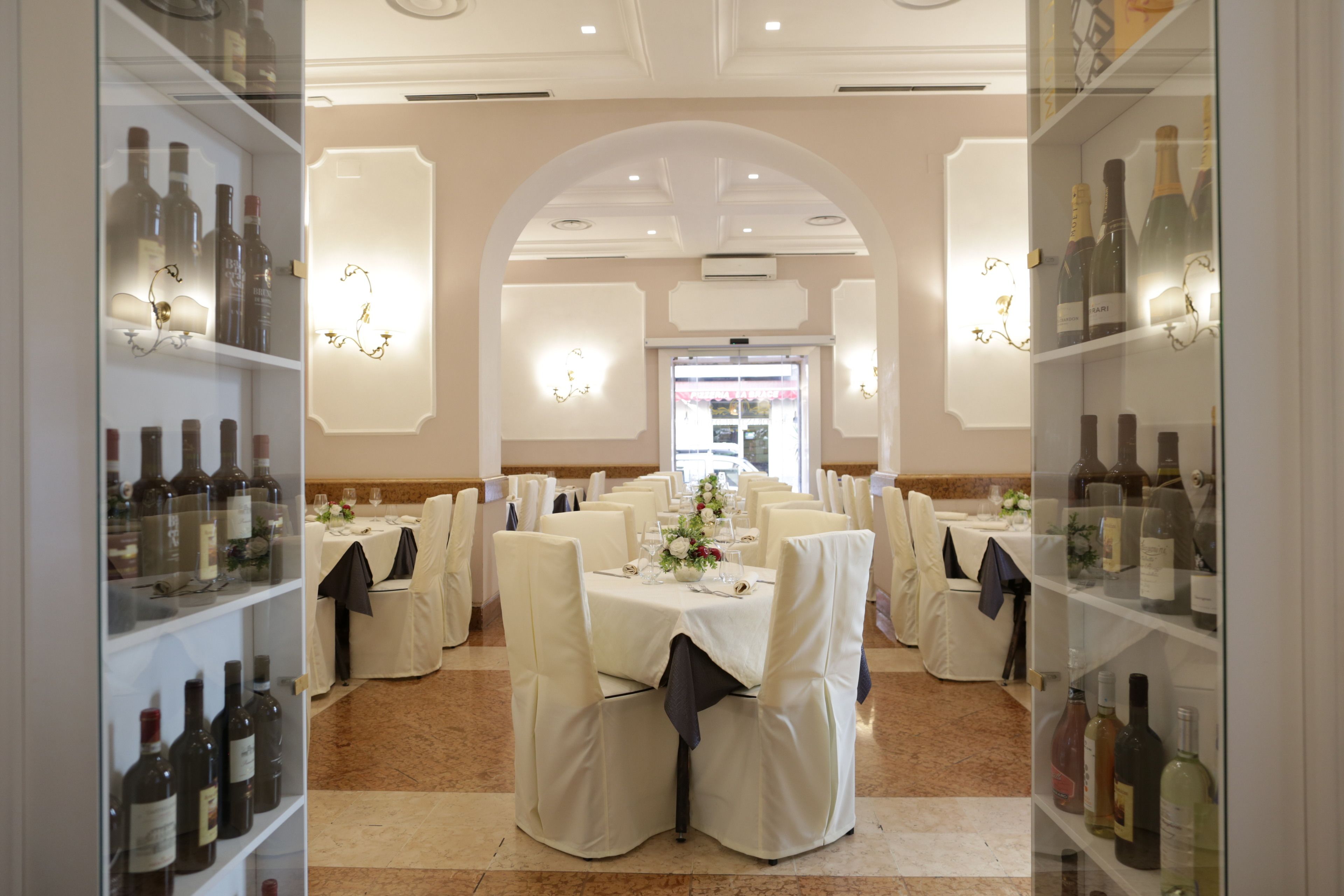 A banquet hall with round tables, white chairs, and wine bottles on shelves.