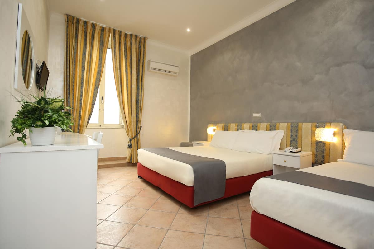 A hotel room with two beds, a desk, a potted plant, and a window with curtains.