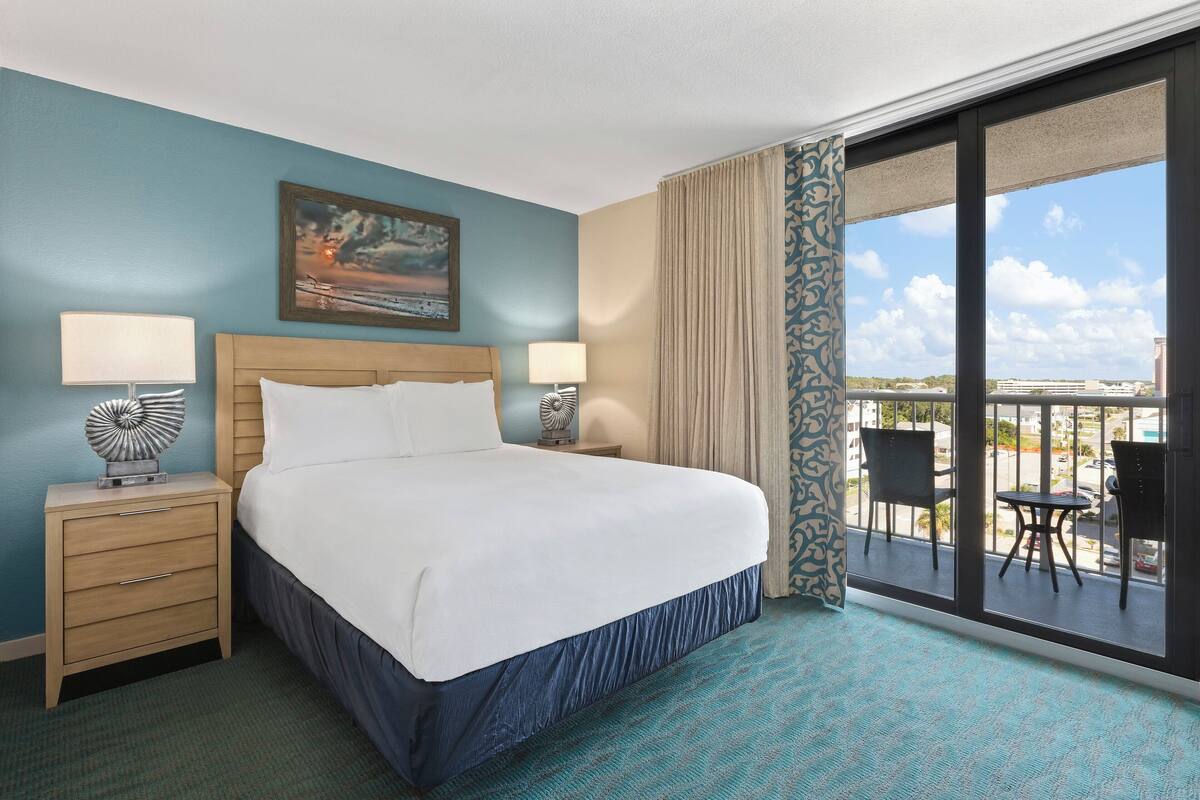 A hotel room with a large bed, two bedside tables with lamps, a painting on the wall, and a balcony with chairs.