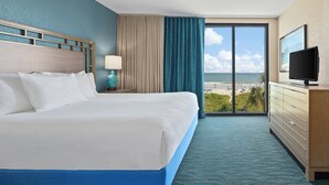A hotel room with a large bed, a TV, a dresser, and a view of the beach.