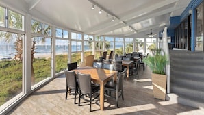 A spacious outdoor dining area with large windows overlooking a beach, furnished with wooden tables and chairs.