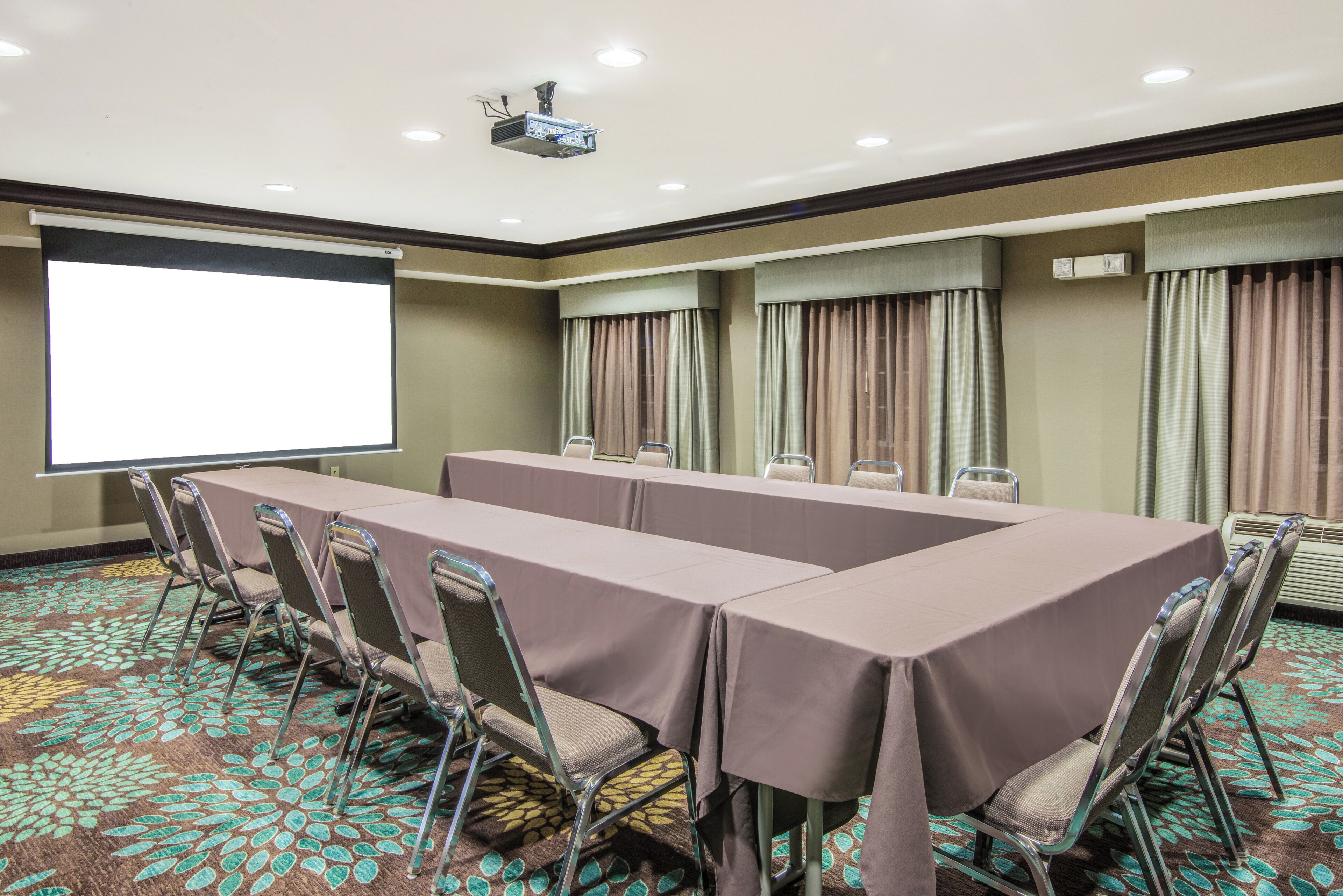 Un salón de conferencias con una larga mesa, sillas, una pantalla de proyector y piso alfombrado.