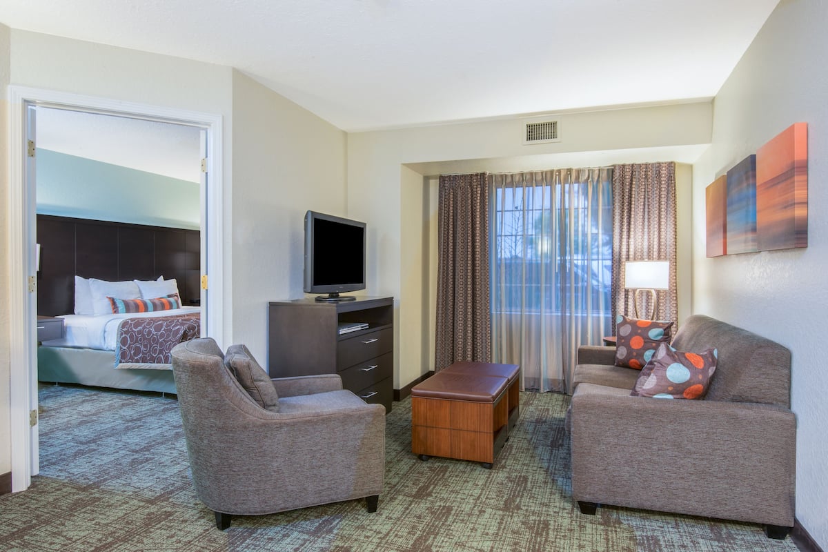 Une chambre d’hôtel comprenant un lit, deux fauteuils, une table basse, un téléviseur et une fenêtre avec des rideaux.