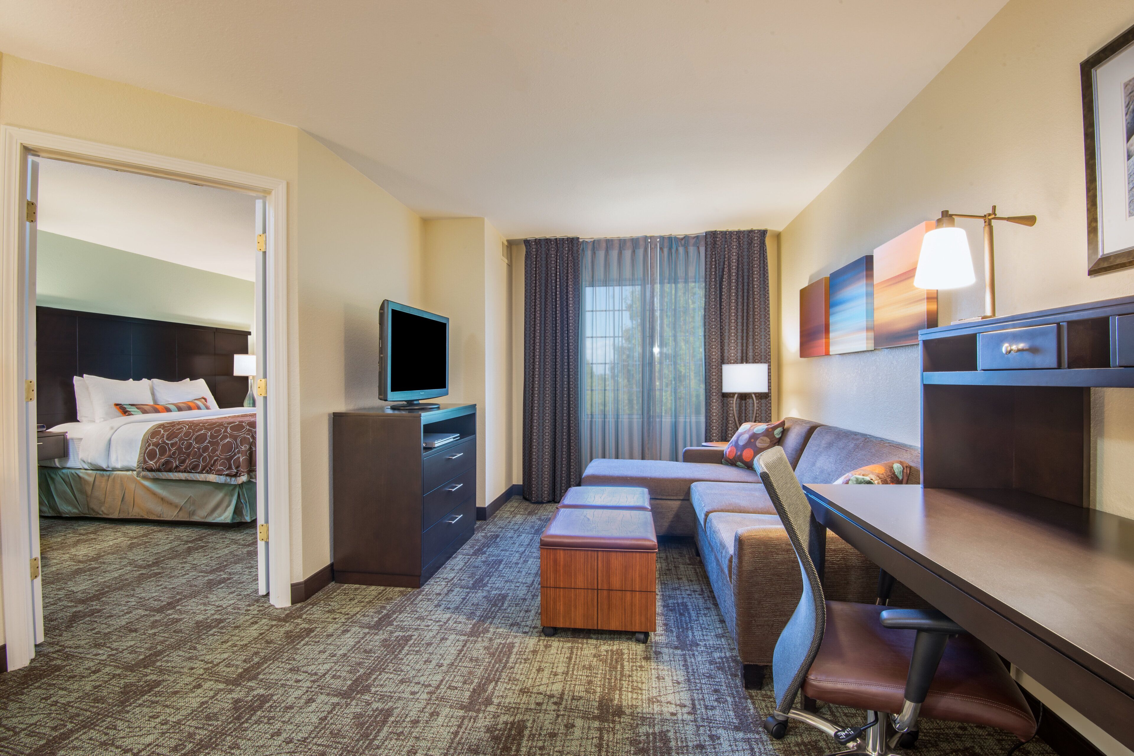 A hotel room with a bed, a desk, a television, a sofa, and a large window with curtains.