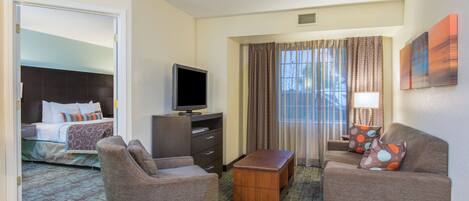 Quarto de hotel com cama, televisão, escrivaninha, duas poltronas, mesa de centro e janela com cortinas.