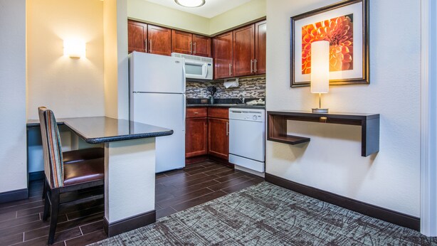 A compact kitchen with dark wood cabinets, a white refrigerator, a microwave, and a dishwasher. There is a small dining table with a chair, a wall-mounted shelf, and a framed picture above the shelf.