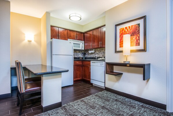 A compact kitchen with dark wood cabinets, a white refrigerator, a microwave, and a dishwasher. There is a small dining table with a chair, a wall-mounted shelf, and a framed picture above the shelf. - Staybridge Suites Allentown West by IHG (Allentown)