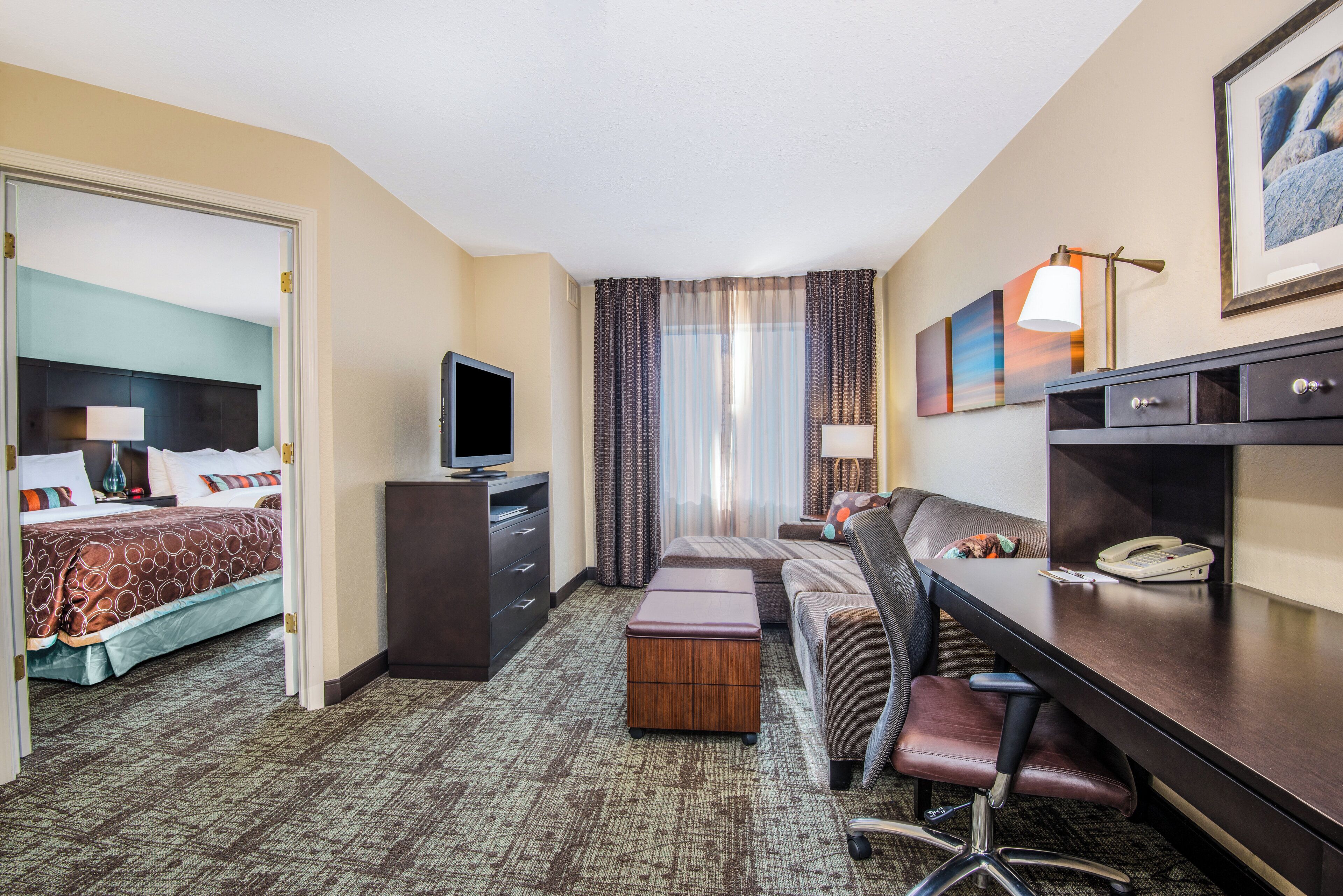 Habitación de hotel con cama, escritorio, silla, televisión, teléfono y ventana con cortinas.