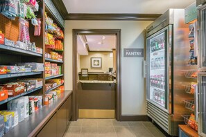 A pantry area with shelves stocked with snacks and beverages, a refrigerator, and a door leading to a kitchen. - Staybridge Suites Allentown West by IHG (Allentown)