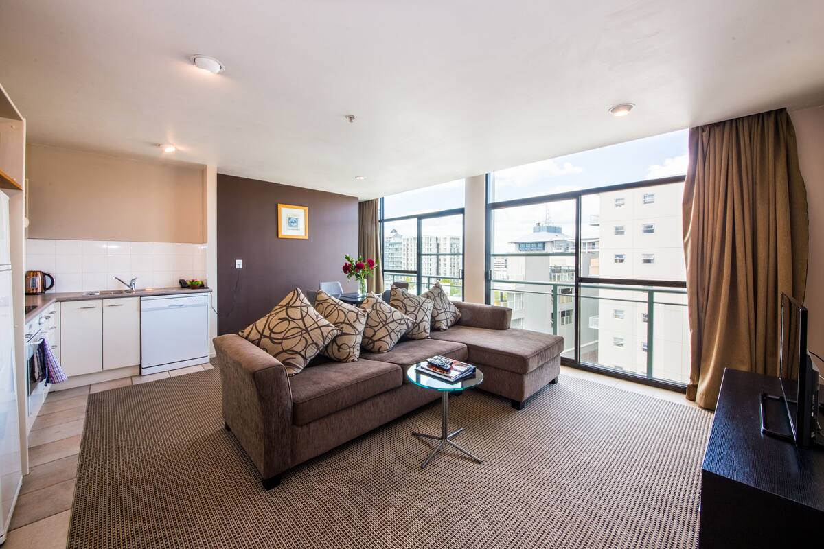 Una sala de estar moderna con un sofá, una mesita de centro y un mueble para televisión. Cuenta con una zona de cocina con lavavajillas y encimera. La habitación tiene ventanas grandes con vistas a edificios.