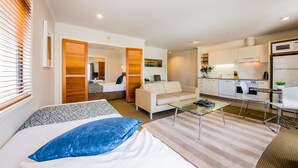 A modern hotel room with a bed, a sofa, a glass coffee table, a kitchen area with a microwave, and a dining table with chairs. - Quest On Nelson (Auckland)