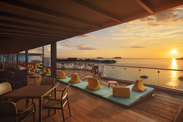 Una terraza con asientos y mesas, con vista a un cuerpo de agua durante el atardecer.