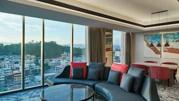 A modern hotel room with a curved sofa, a glass coffee table, and a city view through large windows.