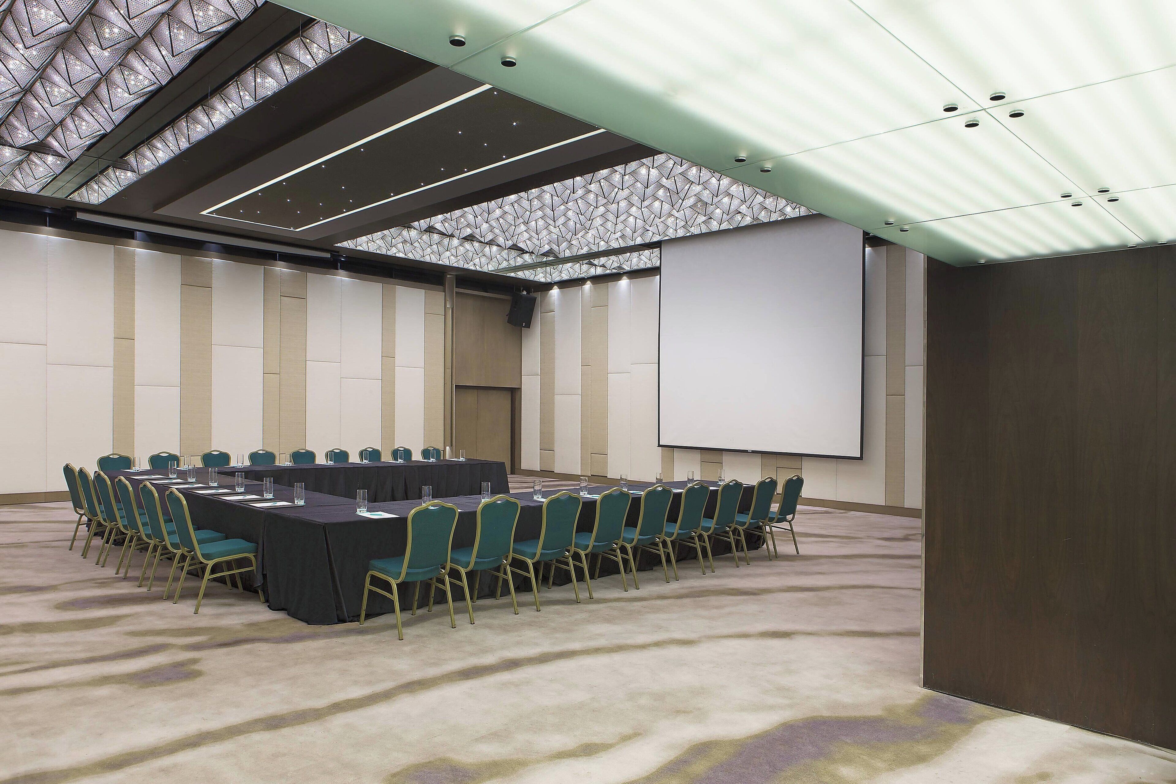 Un salón de conferencias con una larga mesa, sillas, una pantalla de proyector y suelo alfombrado.