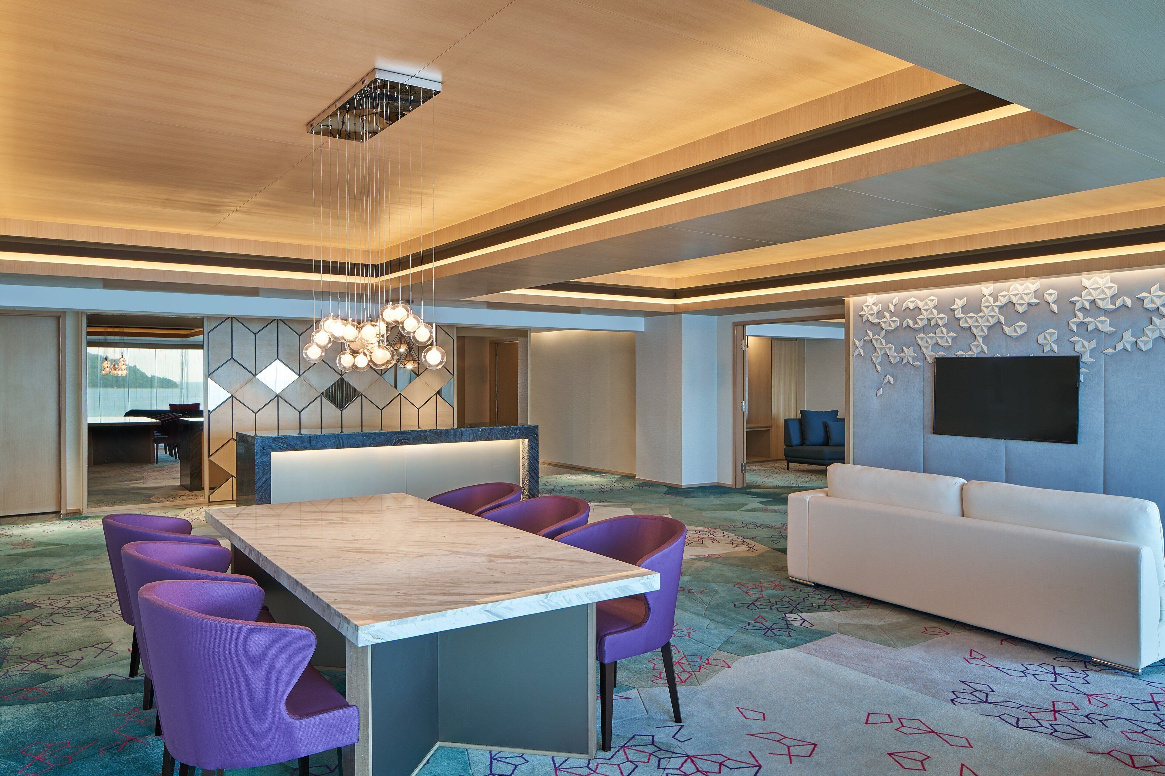 A modern hotel lobby with a wooden reception desk, purple chairs, a white sofa, and a large TV on the wall.