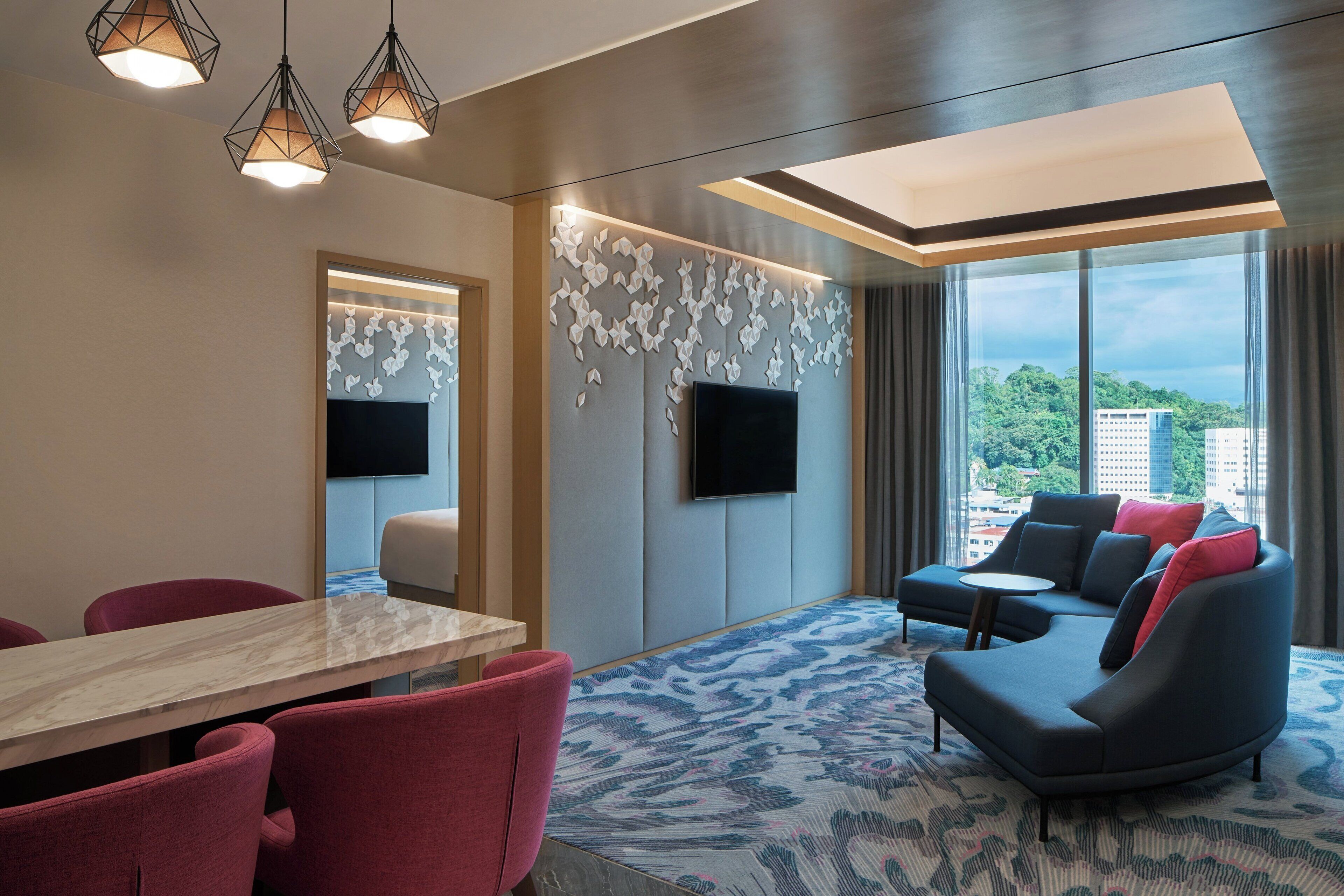 Een hotelkamer met een groot raam, een bank, een televisie, een eettafel en rode stoelen.
