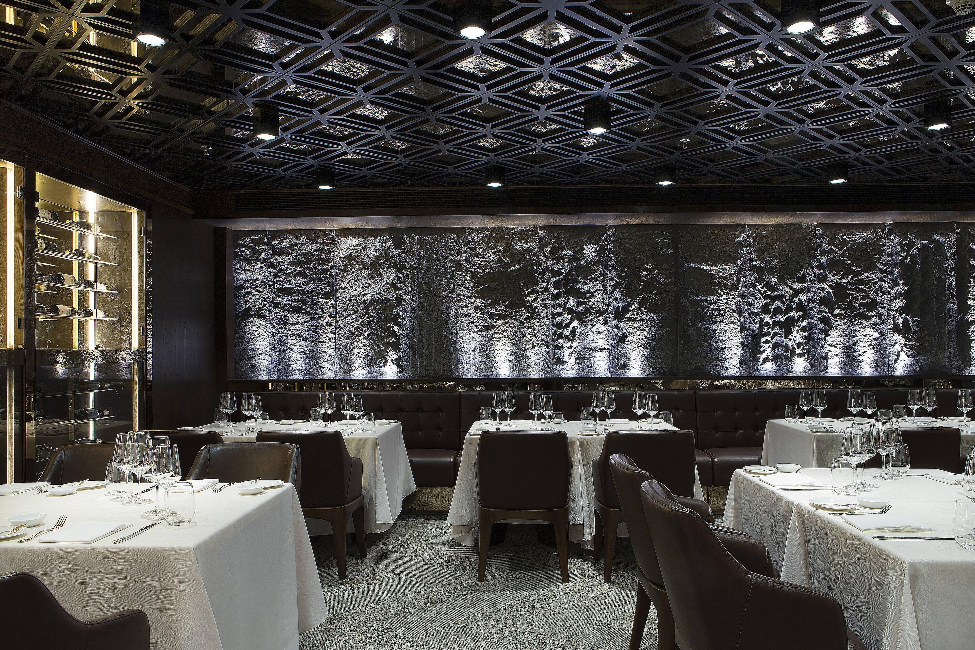 A restaurant with tables set for dining, a large illuminated wall, and a ceiling with a geometric pattern.