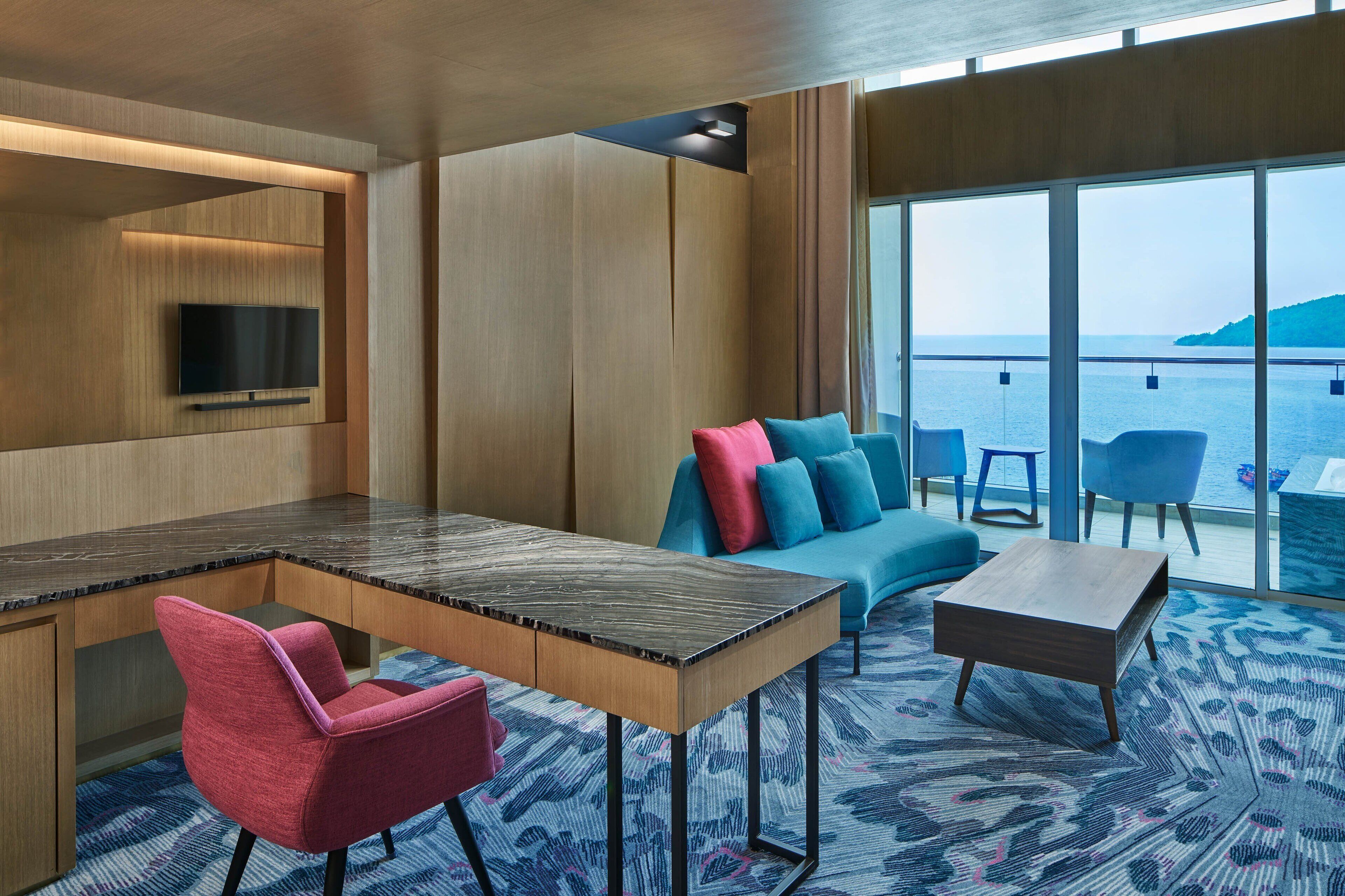 A modern room with a wooden desk, a TV mounted on the wall, a sofa with colorful cushions, and a view of the sea through large windows.