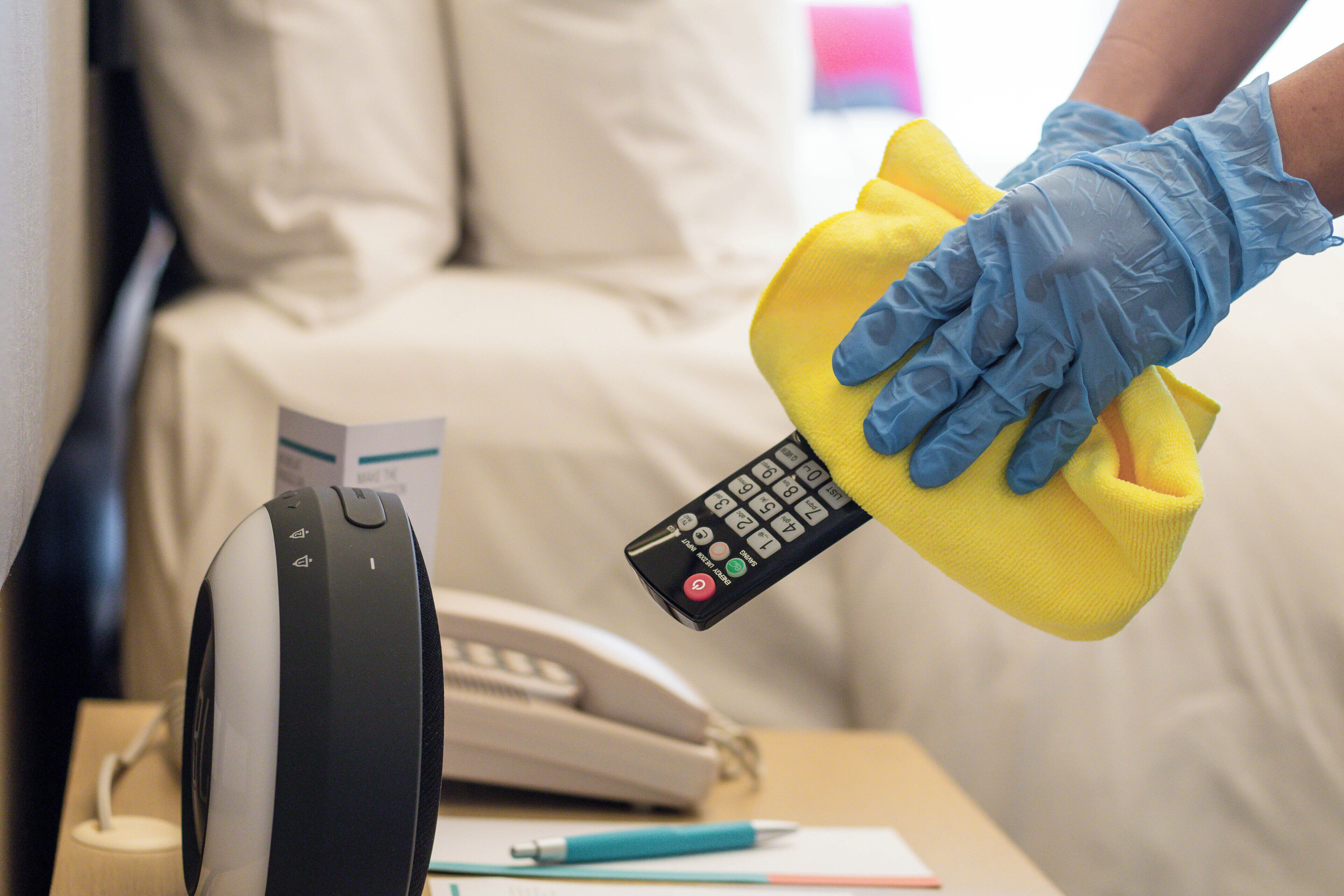 A hand holding a yellow cloth with a remote control, a telephone, and a pen on a table.