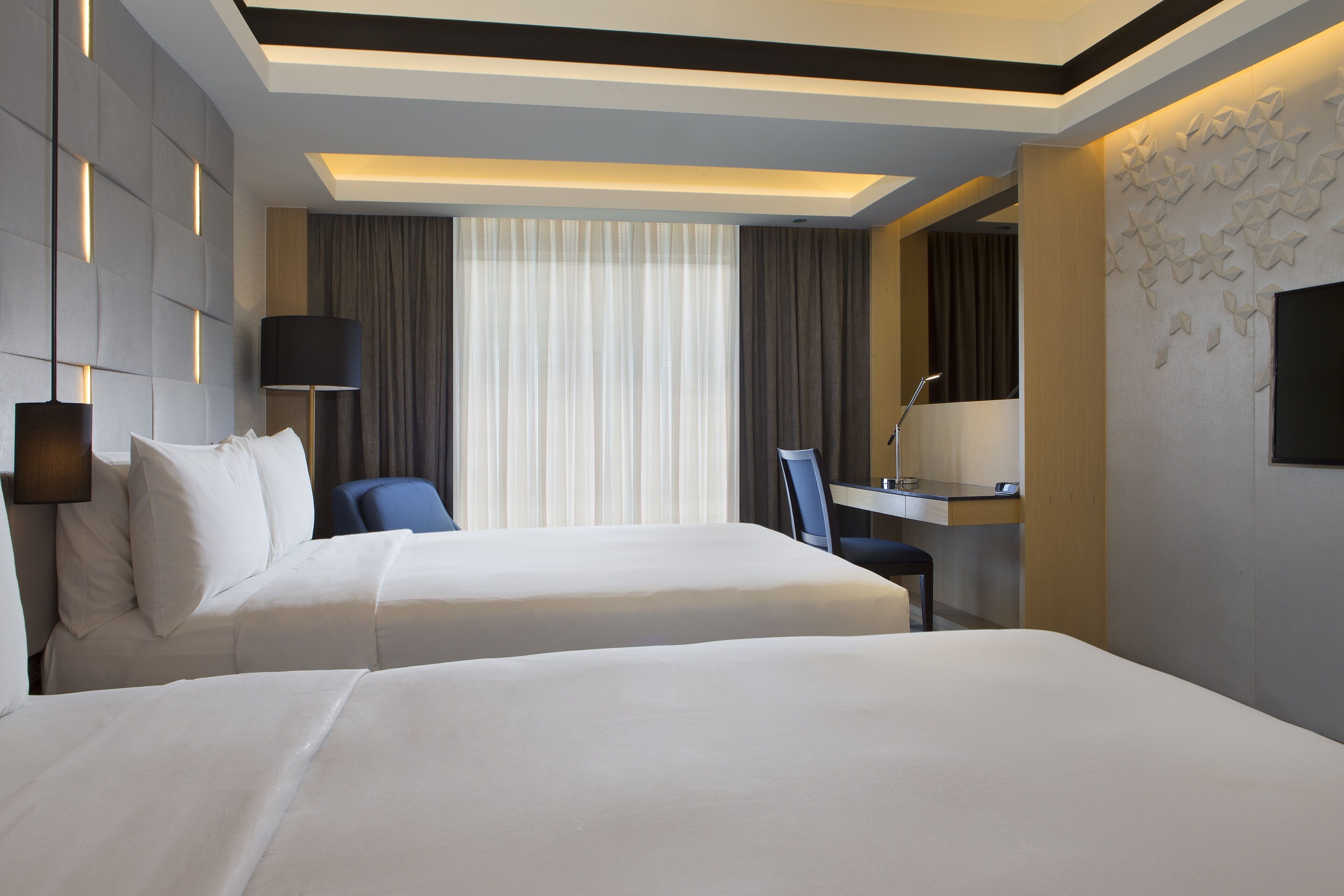 Habitación de hotel con dos camas, un escritorio, una silla y un ventanal con cortinas.