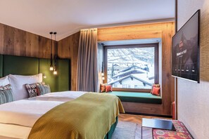 Une chambre d’hôtel moderne avec un grand lit, un banc recouvert d’un tissu vert placé près de la fenêtre, et une vue sur une montagne enneigée.