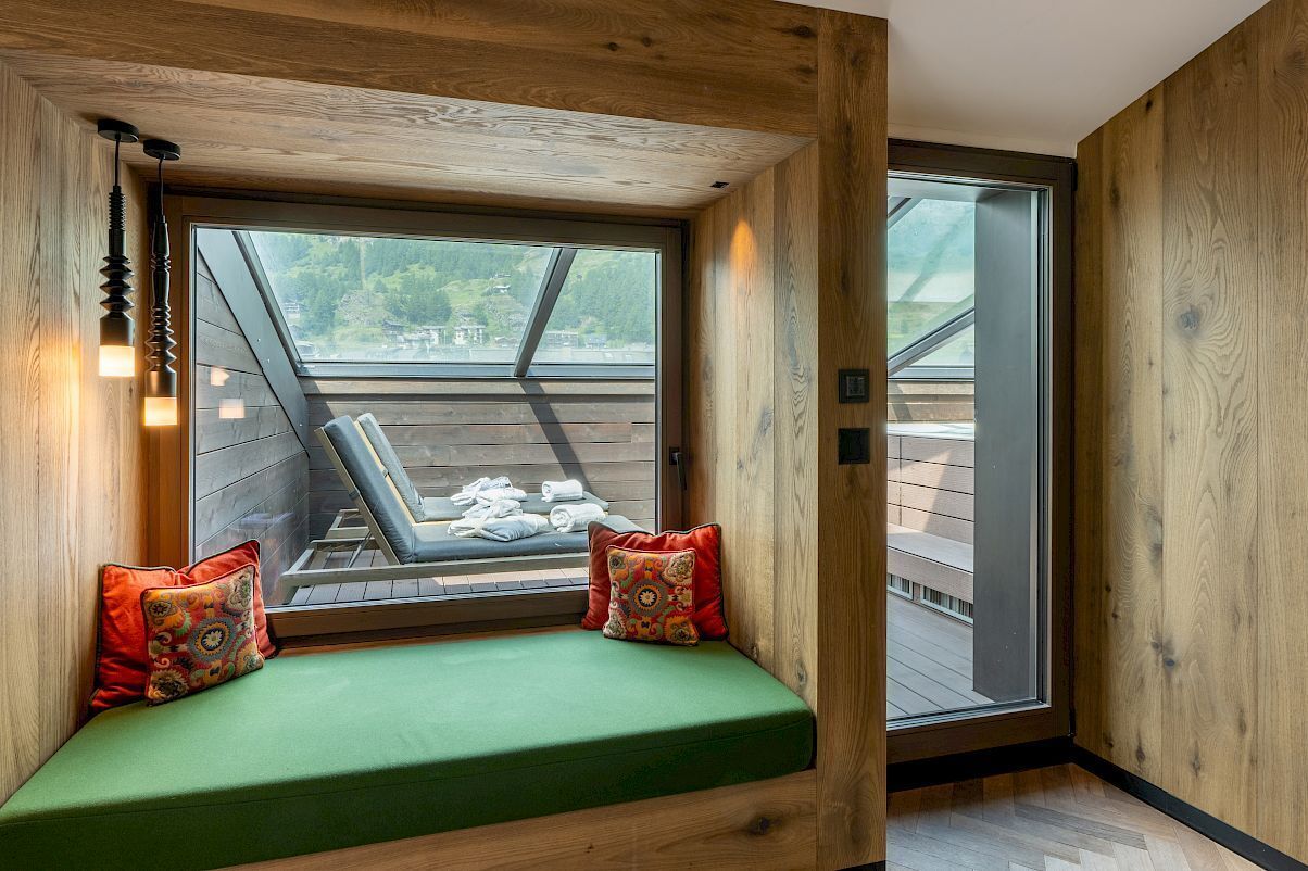 Una habitación acogedora con detalles de madera, un banco tapizado en verde con cojines de colores y un amplio ventanal con vistas a una terraza amueblada.