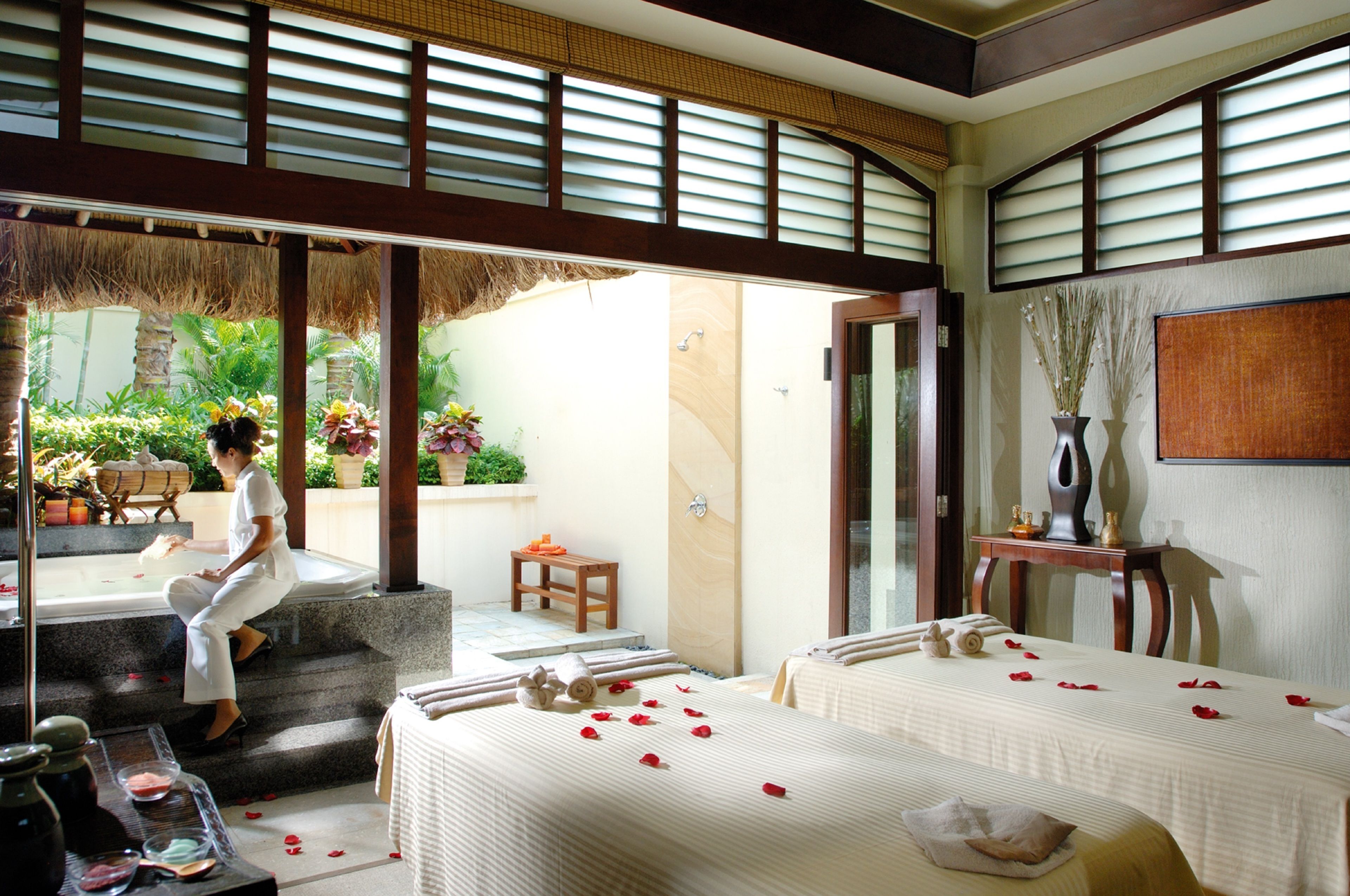 A hotel room with a balcony, a bed with rose petals, a small table with a vase, and a view of the garden.