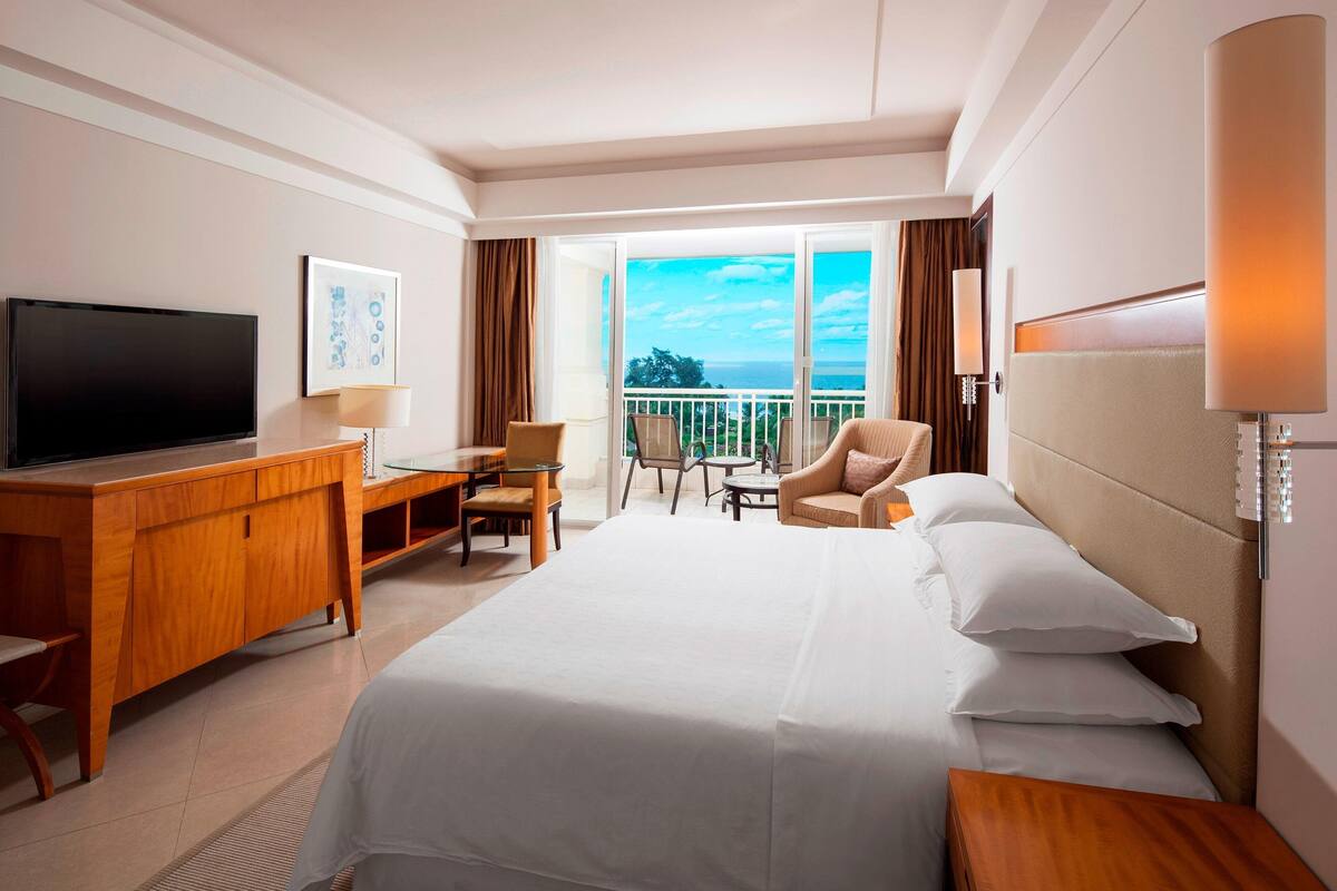 Habitación de hotel con cama, escritorio, televisor, silla y balcón con vistas.