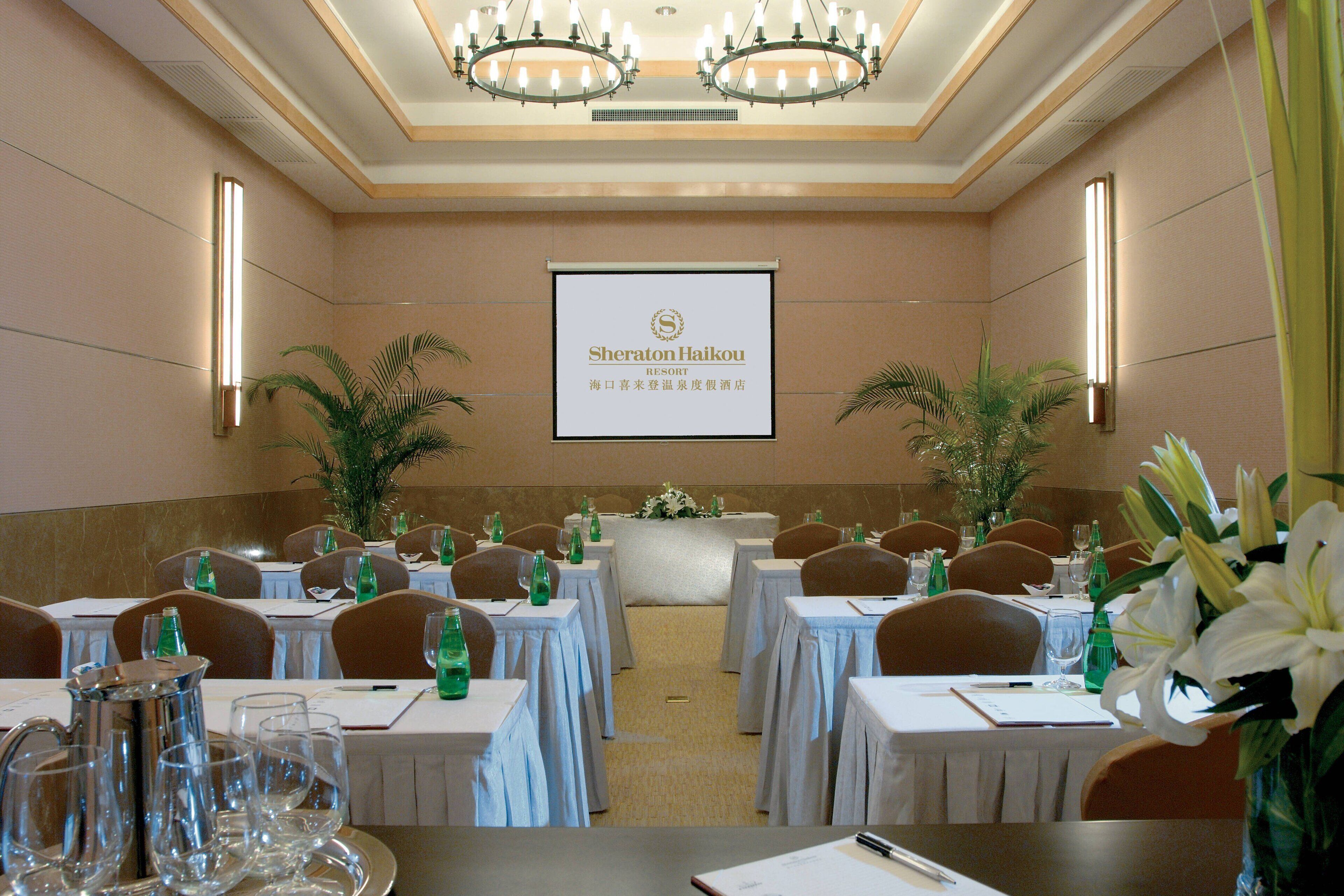 Uma sala de conferências no Sheraton Haikou Resort, com mesas preparadas para uma reunião, arranjos florais e uma tela de projetor exibindo o nome do resort.