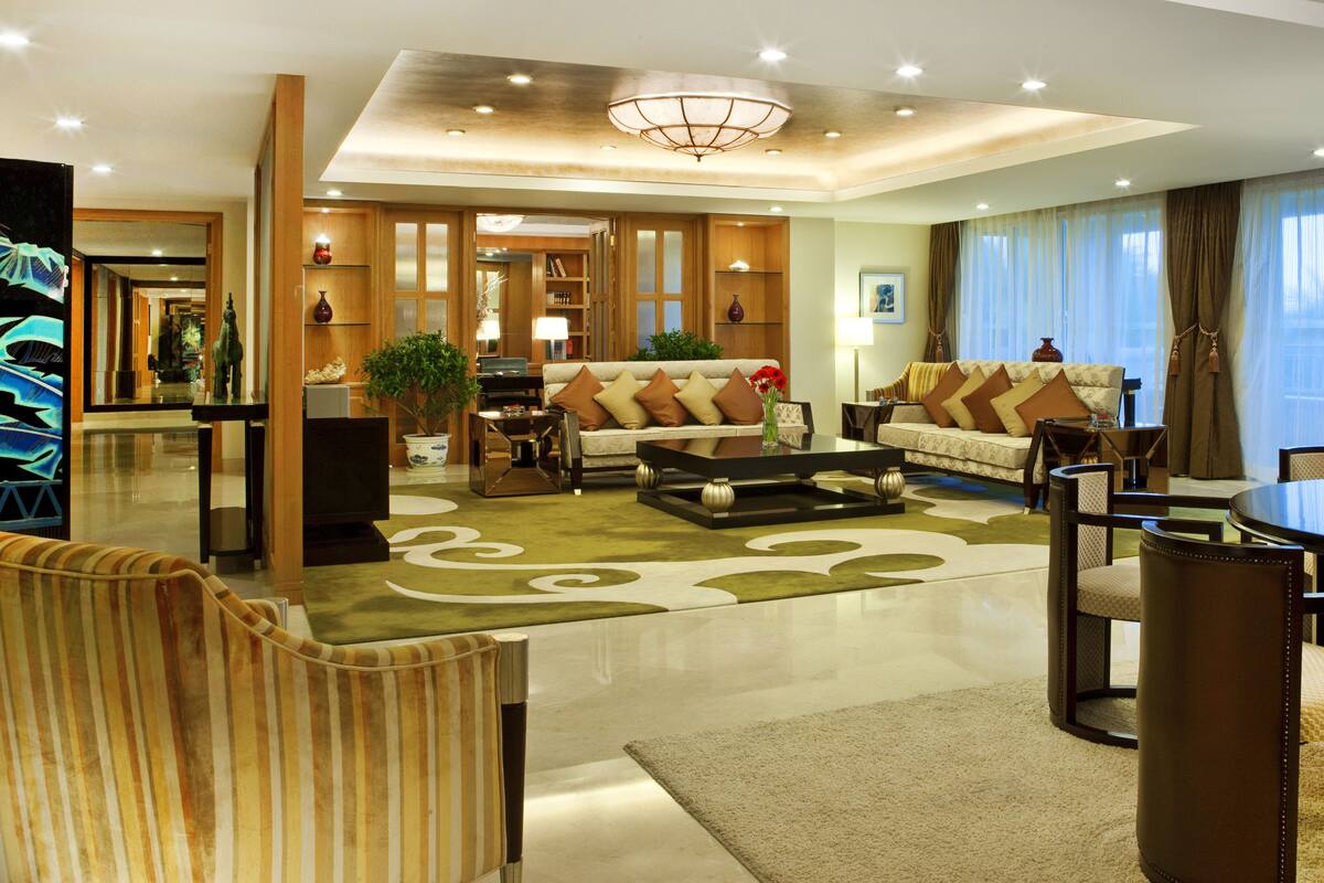 Un amplio vestíbulo de hotel con diseño moderno, que cuenta con una zona de estar central con sofás y una mesa de comedor, rodeado por un espejo grande y plantas decorativas.