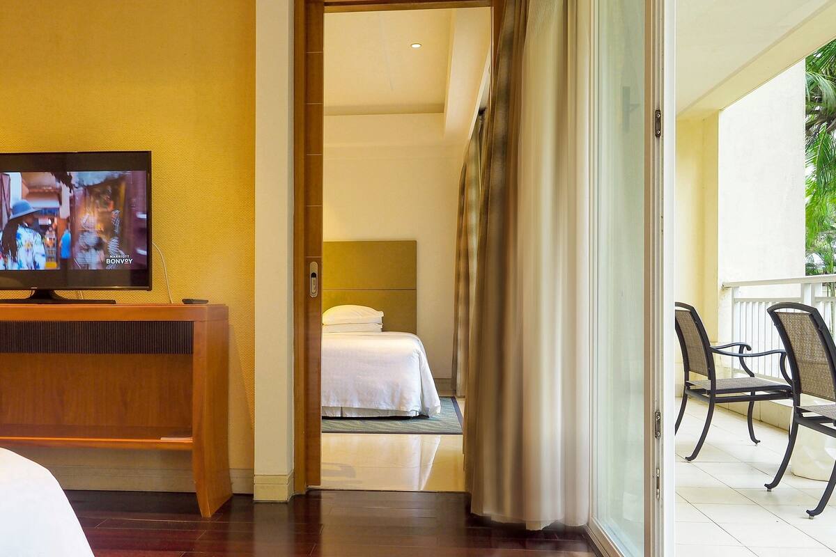 Habitación de hotel con cama, televisor sobre un mueble de madera y balcón con sillas.