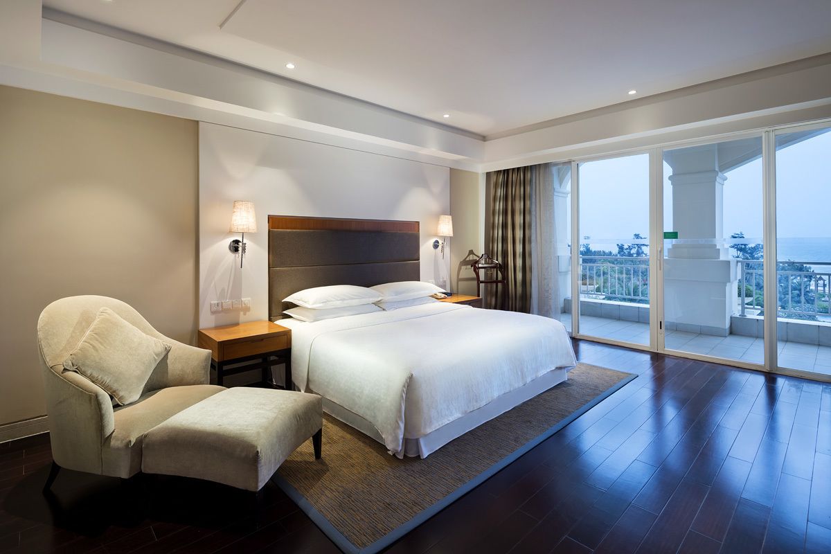 A hotel room with a large bed, a chair, a nightstand, and a view of the sea through large windows.
