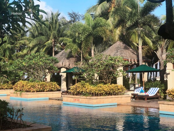 A pool area with lounge chairs, palm trees, and a thatched-roof structure.