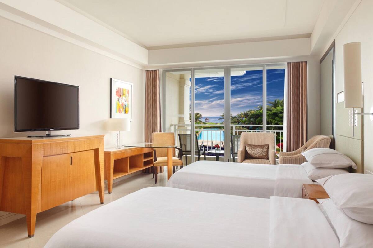 Habitación de hotel con dos camas, un escritorio, un televisor y un balcón con vista al océano.