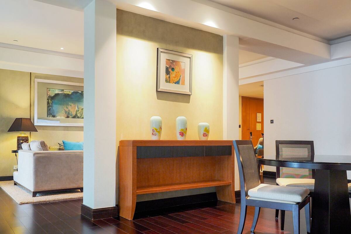 Un hall de hotel con un sofá, una consola de madera con tres jarrones decorativos y un comedor con mesa y sillas.